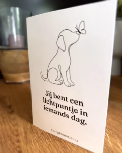 Witte dubbele kaart die rechtop staat op een houten tafel, met een lijntekening van een hondje en een vlinder en de tekst Jij bent een lichtpuntje in iemands dag